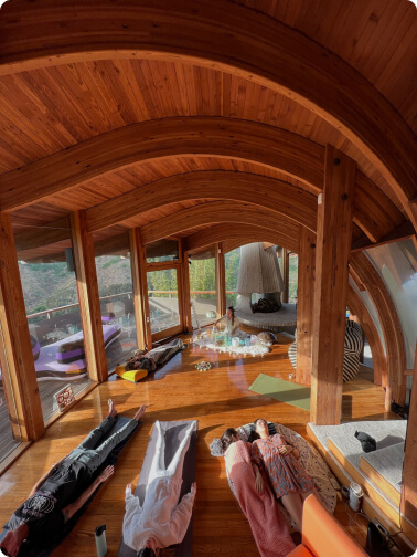 mediation hall with big windows and wooden ceiling with multiple multiple people laying on mats and doing meditation