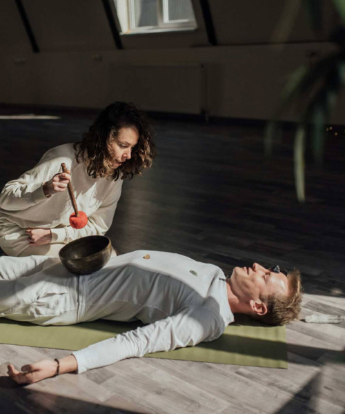 guy lies on his back with a healing sound bawl placed on his stomach and girl helps him do meditation